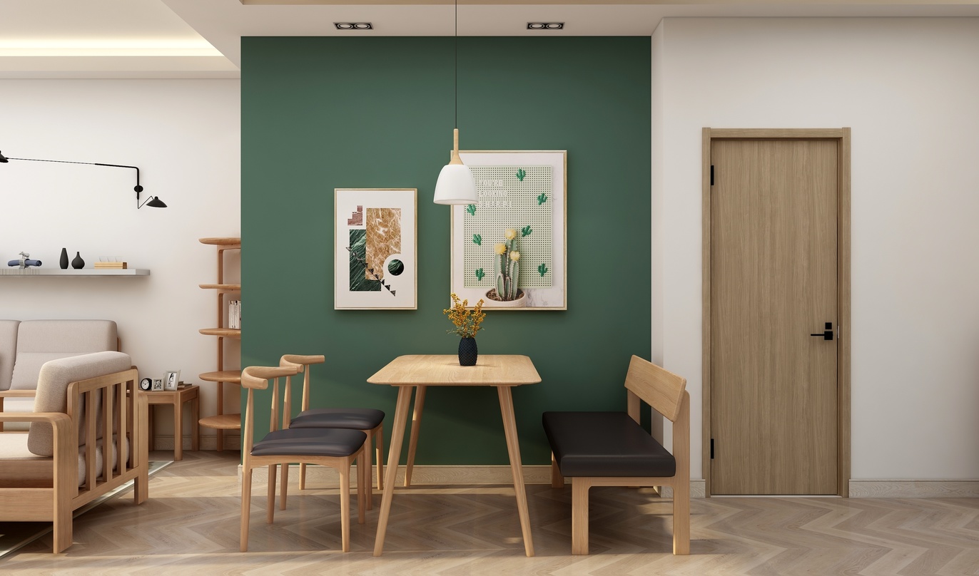 图中是一个餐厅厨房，墙面是墨绿色，地面是浅棕色的木地板。墙面左侧挂着两幅装饰画，下面有一张木质小桌子，桌子两边各放着一把木质椅子，椅子背上都放着黑色的坐垫。桌子左侧角落有一个木质楼梯，直通上一个房间。房间的一角放着一个灰色的沙发，沙发上放着三个白色的靠垫。房间的一侧是门，门是白色的，上面有木框。房间的另一侧是厨房，厨房的台面上铺了白色的小瓷砖，柜子门是白色的，上面的把手是金色的。墙上还挂着一个白色的灯。
